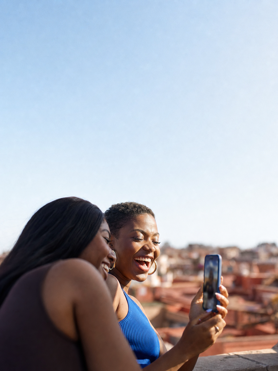 Two friends laughing at a phone on a rooftop above terracotta roofs.